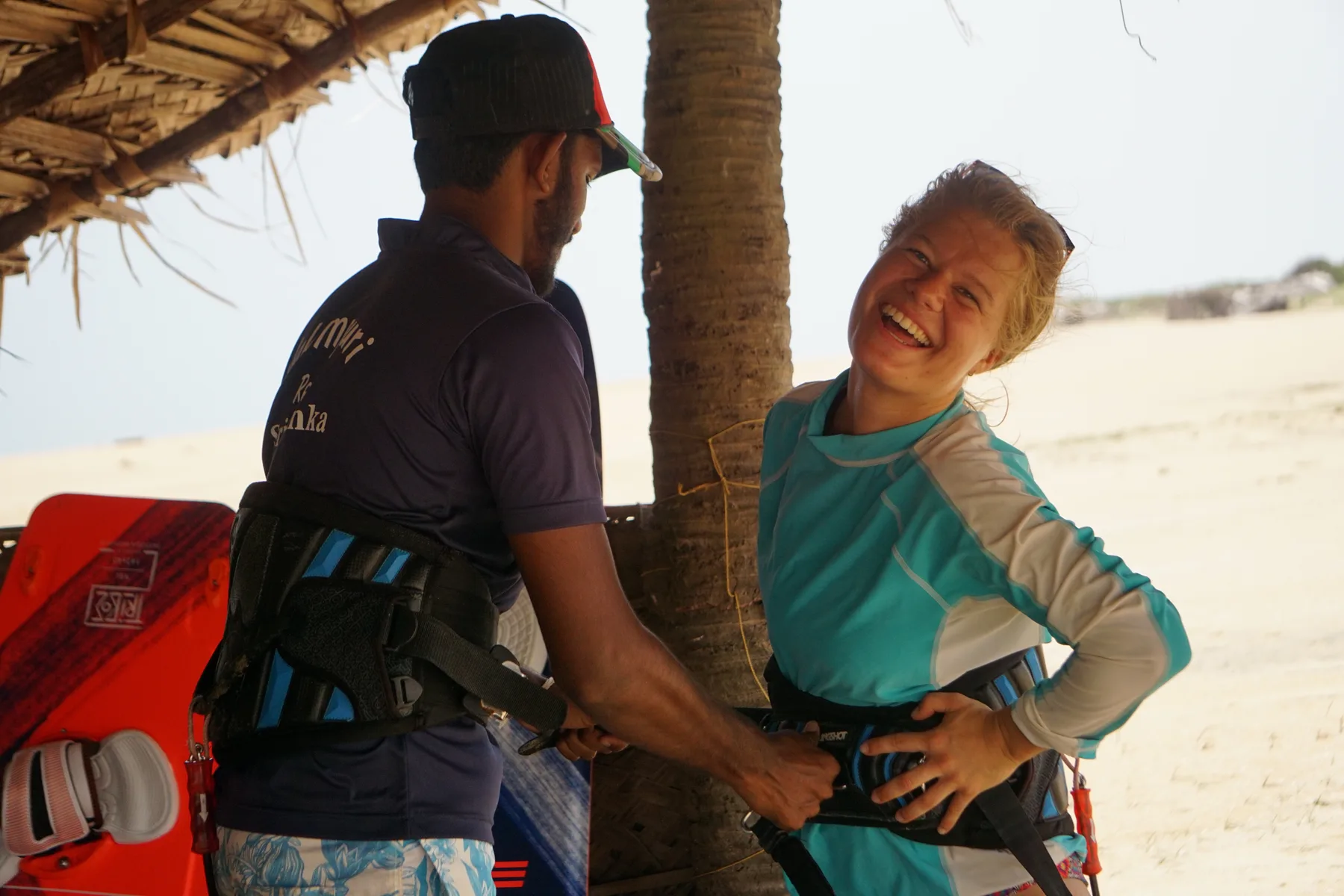 Instructor helping a laughing beginner into a kite harness on the beach at Kalpitiya